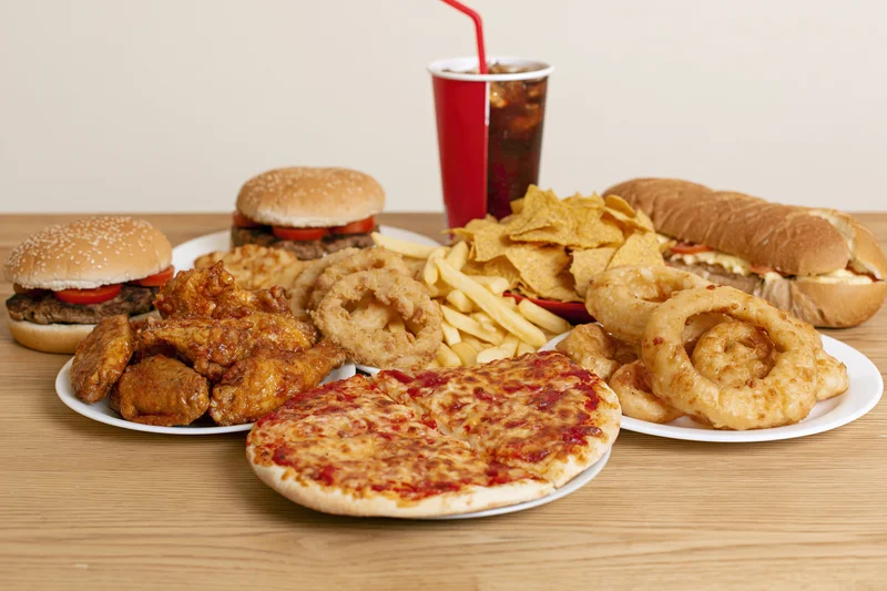 Assorted fast food items arranged on a wooden table including burgers, pizza, fries, onion rings, chicken wings, soda and chips
