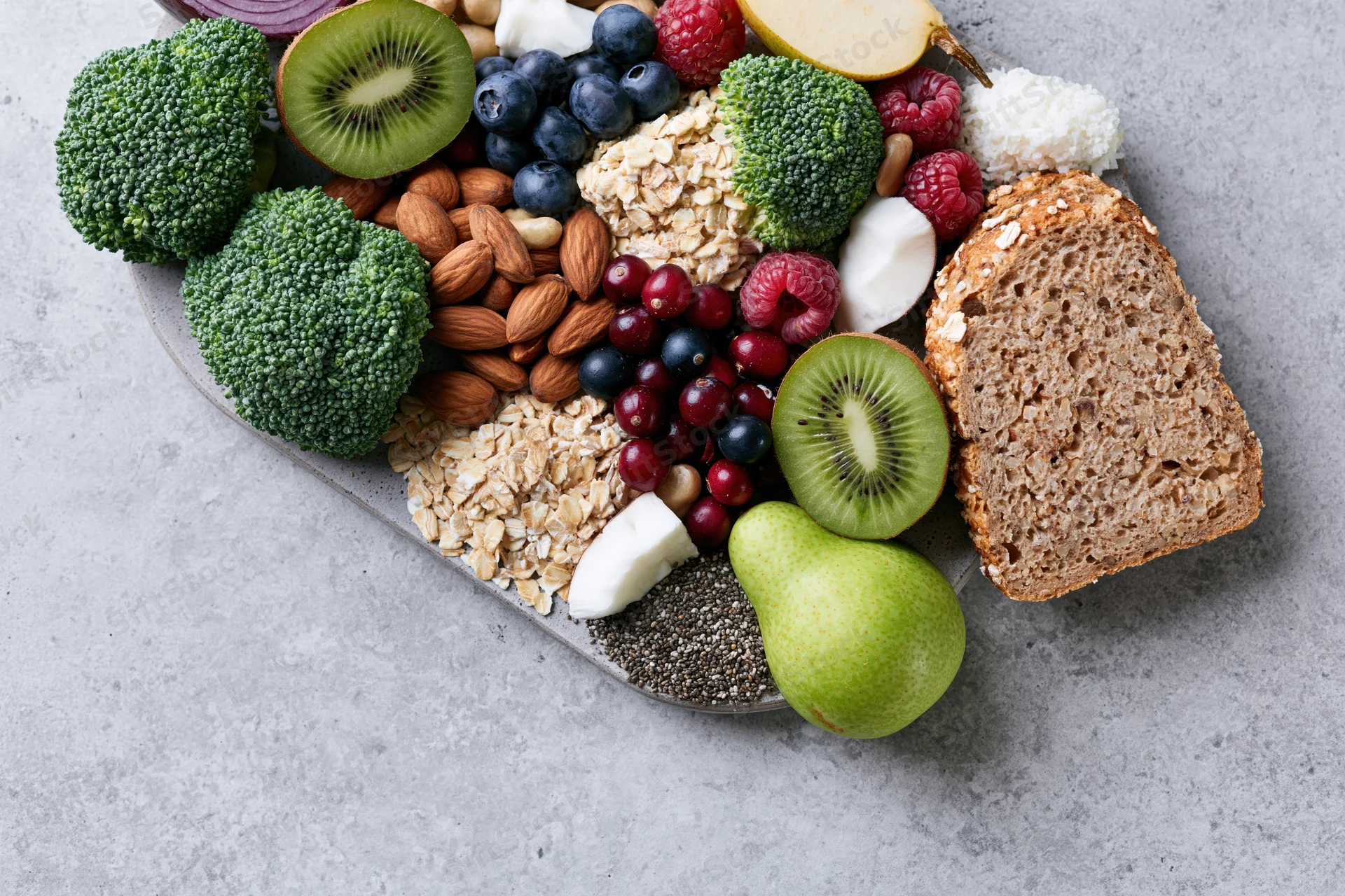 Assorted fresh fruits vegetables nuts seeds and whole grains arranged on a gray surface