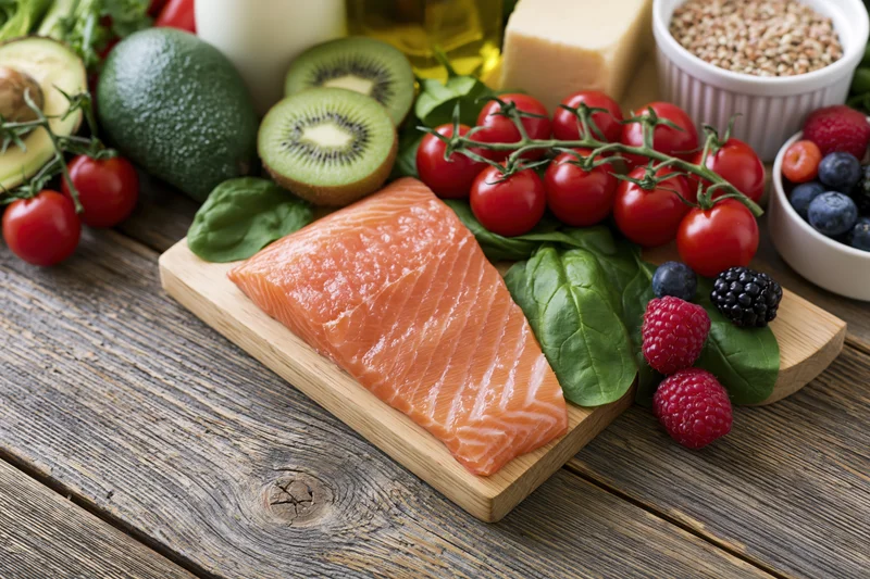 Raw salmon fillet on a wooden cutting board surrounded by fresh fruits vegetables and dairy on a rustic wooden table