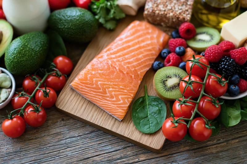 Raw salmon fillet on wooden cutting board surrounded by fresh vegetables fruits and dairy on rustic table