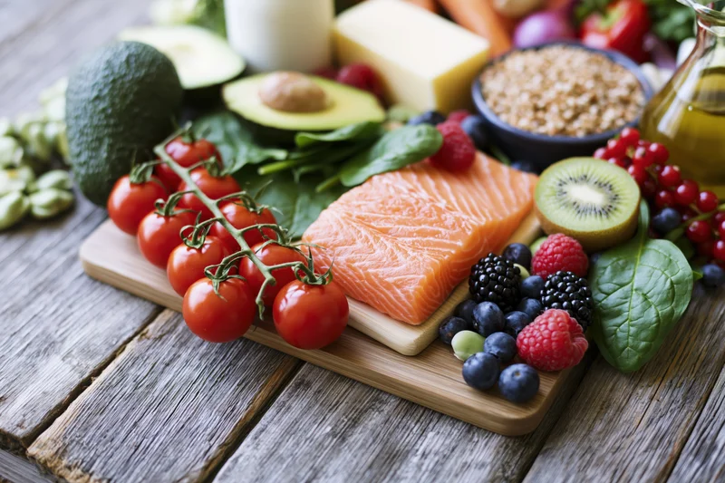 Salmon fillet with tomatoes, avocado, berries and greens arranged on a wooden cutting board with dairy and grains nearby