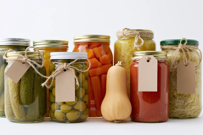 Glass jars filled with pickled vegetables and squash arranged in a row on a white surface