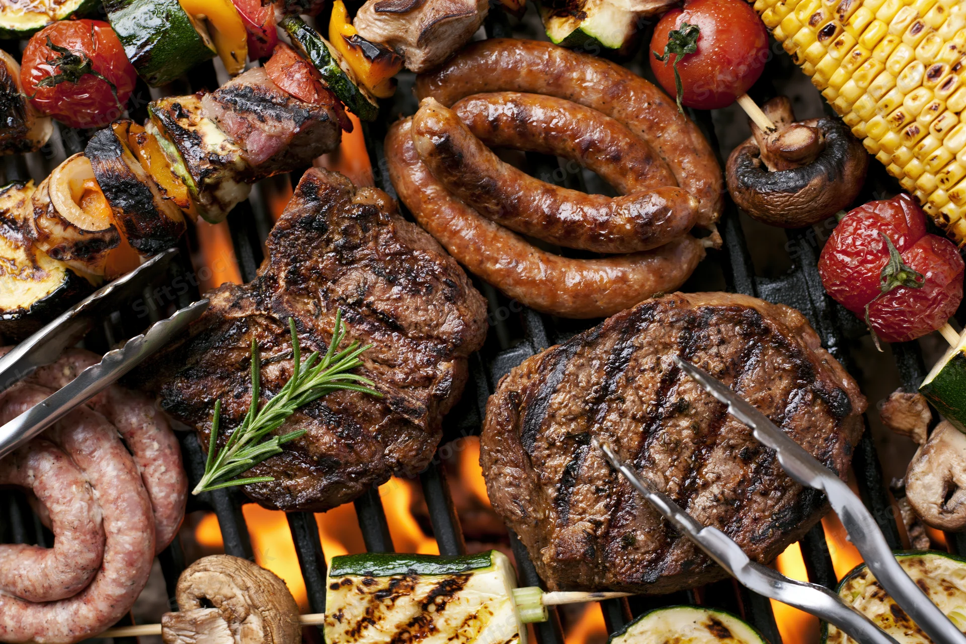 Assorted grilled meats including steaks, sausages, vegetables and corn on a flaming barbecue grill