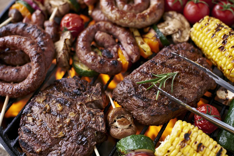 Assorted grilled meats and vegetables cooking on a barbecue grill with flames visible beneath