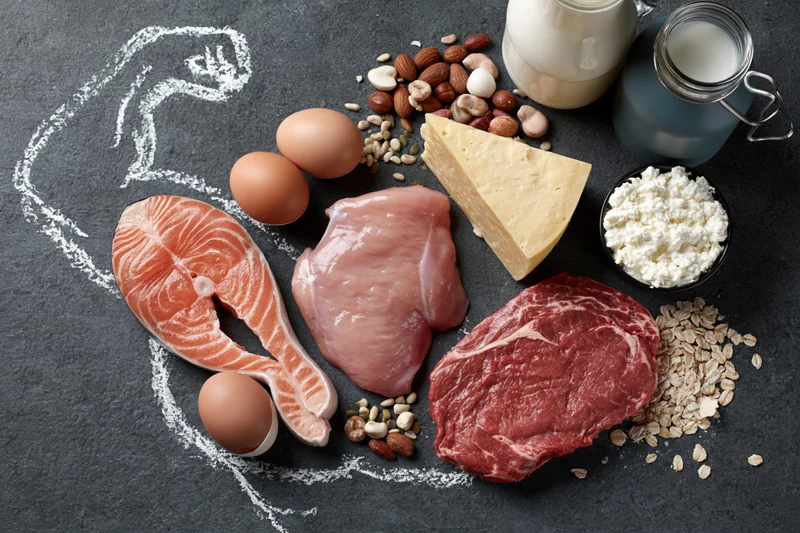 High-protein foods arranged in a flat lay on dark chalkboard surface with chalk outline of a muscular arm