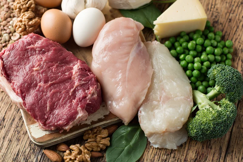 Assorted high-protein foods including meat, chicken, fish, eggs, cheese, broccoli, peas, and nuts arranged on a rustic wooden table