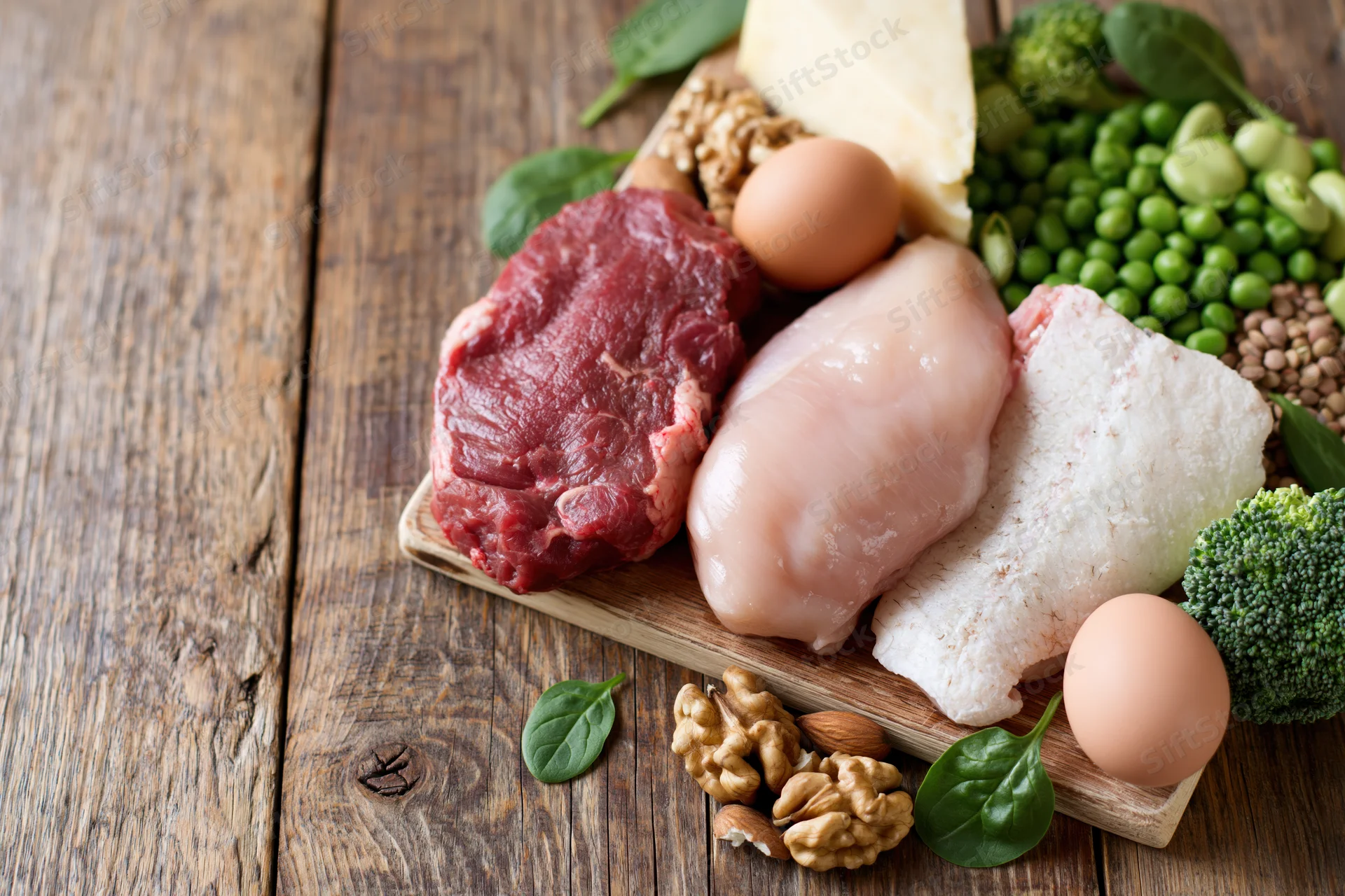 Assorted high-protein foods including meat, eggs, fish, vegetables and nuts arranged on a rustic wooden cutting board
