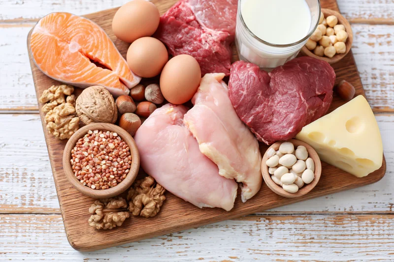 High-protein foods arranged on a wooden cutting board with salmon, beef, chicken, eggs, milk, cheese, nuts and beans