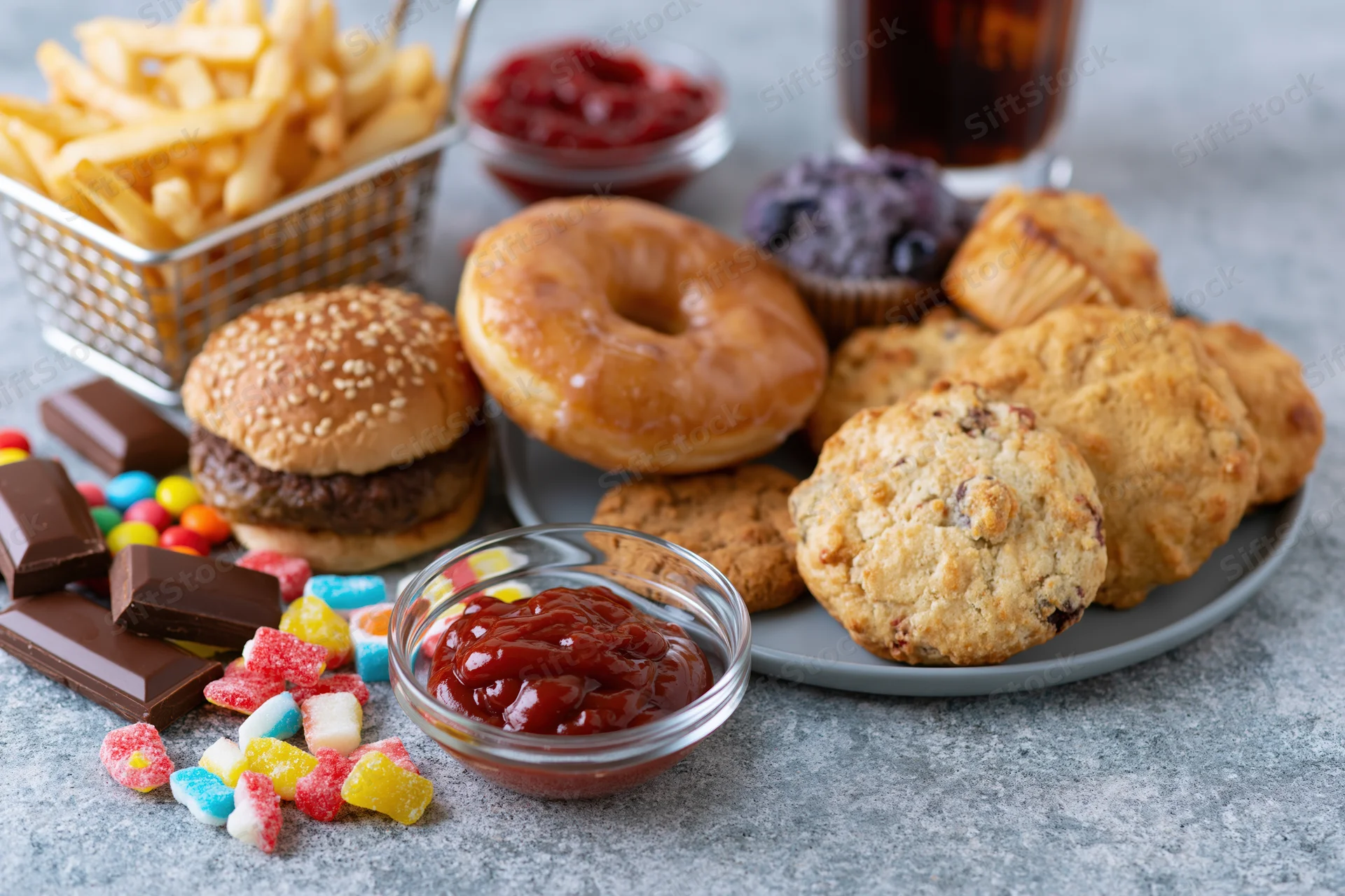 Assorted junk food including burger, donut, fries, cookies, candy and soda arranged on a grey concrete surface