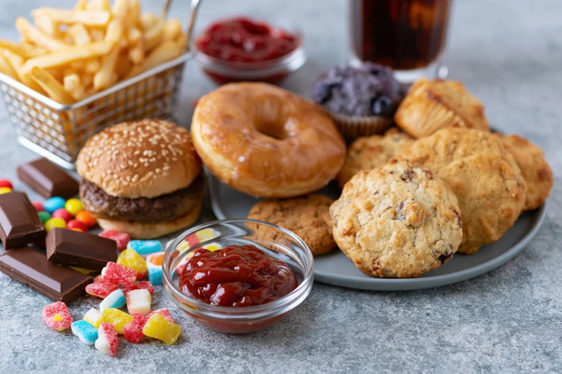 Assorted junk food including burger, donut, fries, cookies, candy and soda arranged on a grey concrete surface