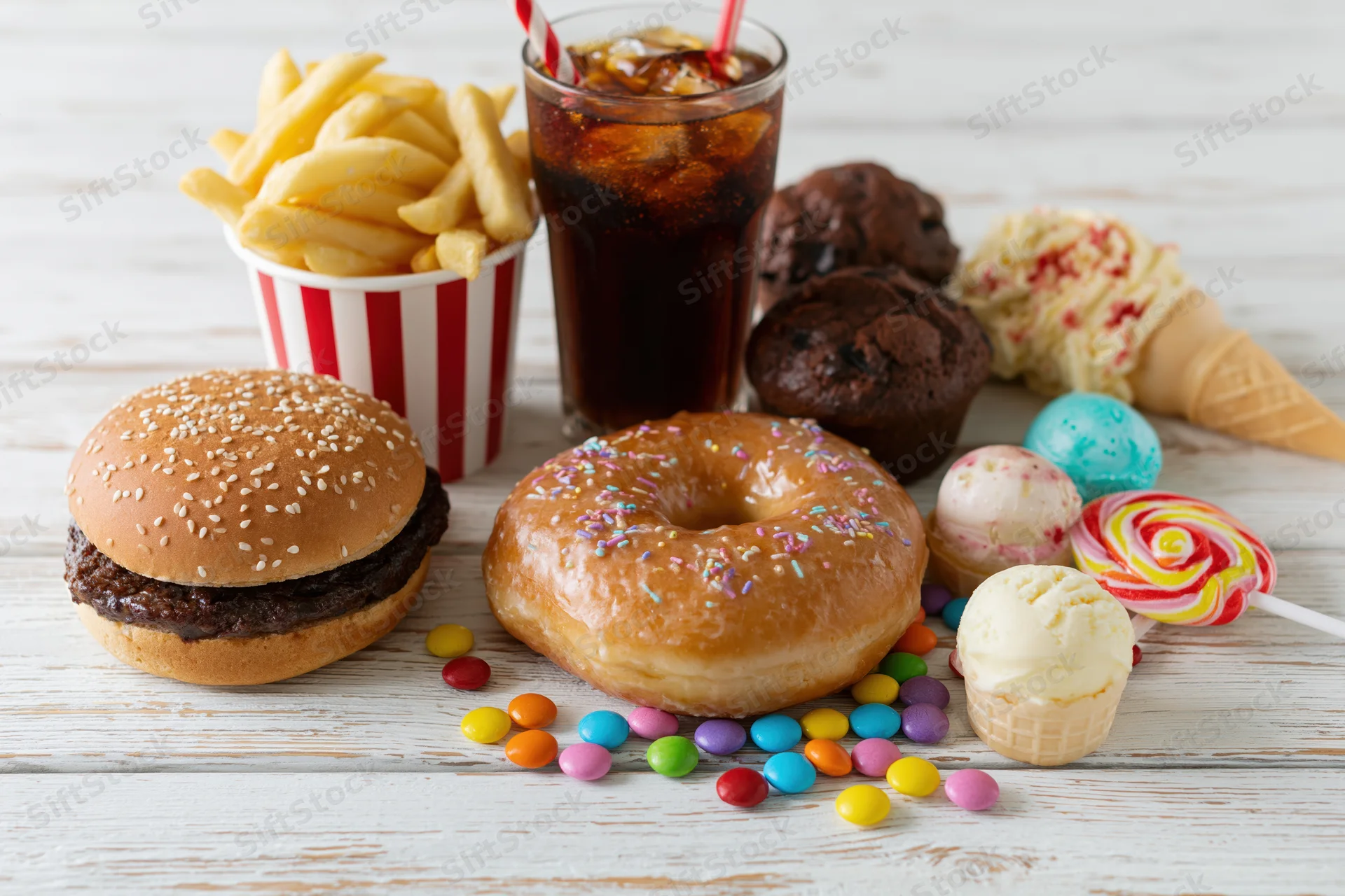 Assorted junk food including burger, donut, fries, soda, ice cream and candy arranged on white wooden surface