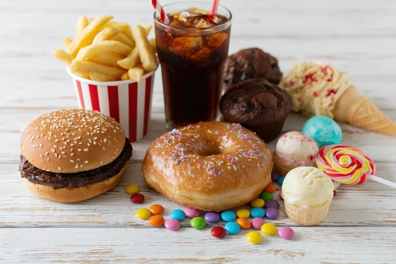 Assorted junk food including burger, donut, fries, soda, ice cream and candy arranged on white wooden surface