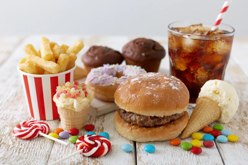 Assorted junk food including burger, fries, soda, ice cream and candy arranged on a white wooden table
