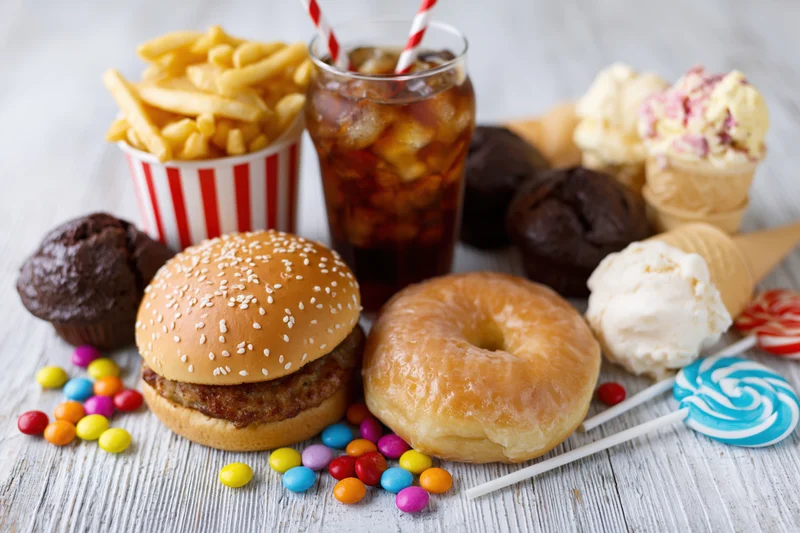 Assorted junk food including hamburger, donut, fries, soda, ice cream and candy arranged on a white wooden tabletop