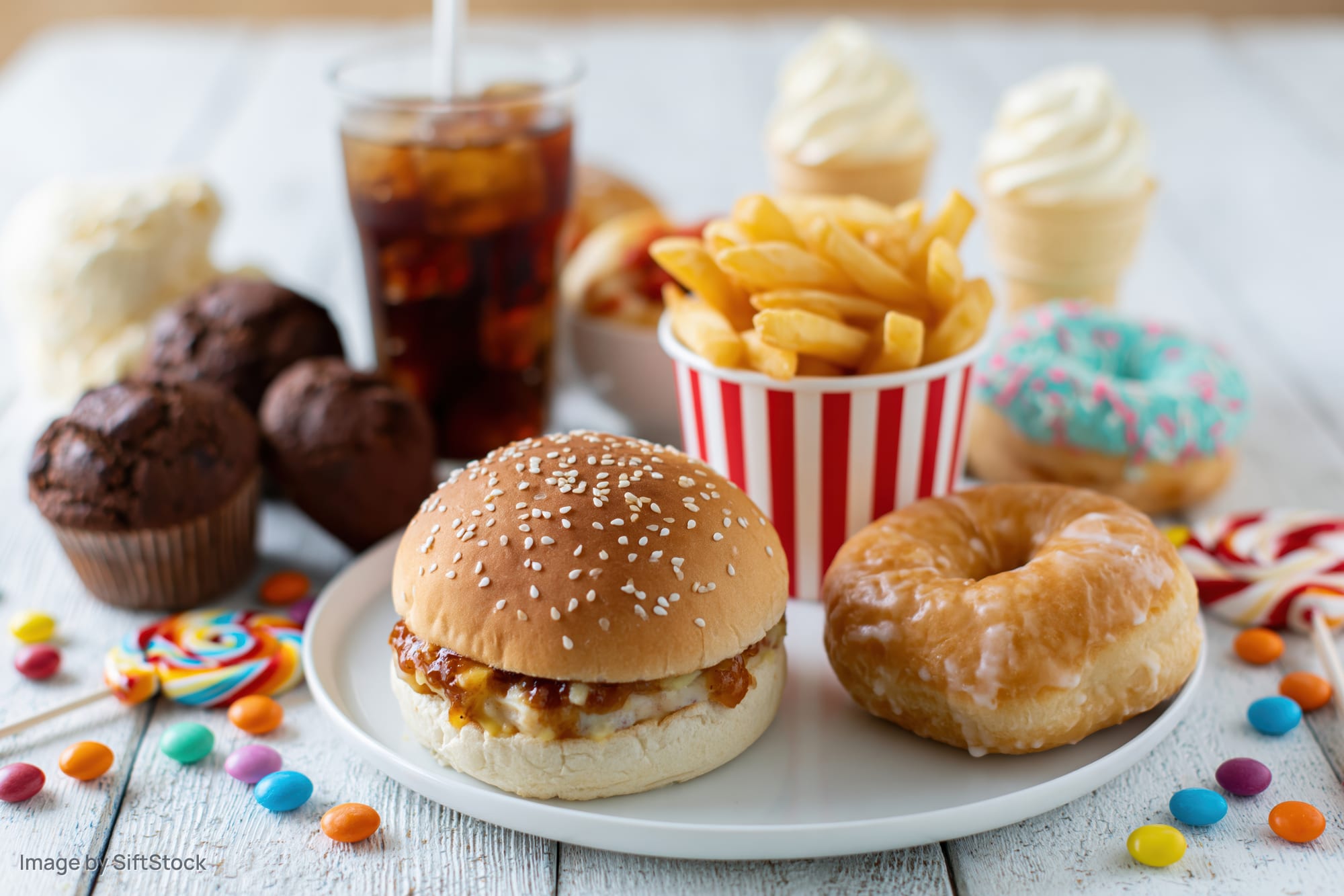 Assorted junk food including burger, donut, fries, soda, and candy arranged on a white wooden table