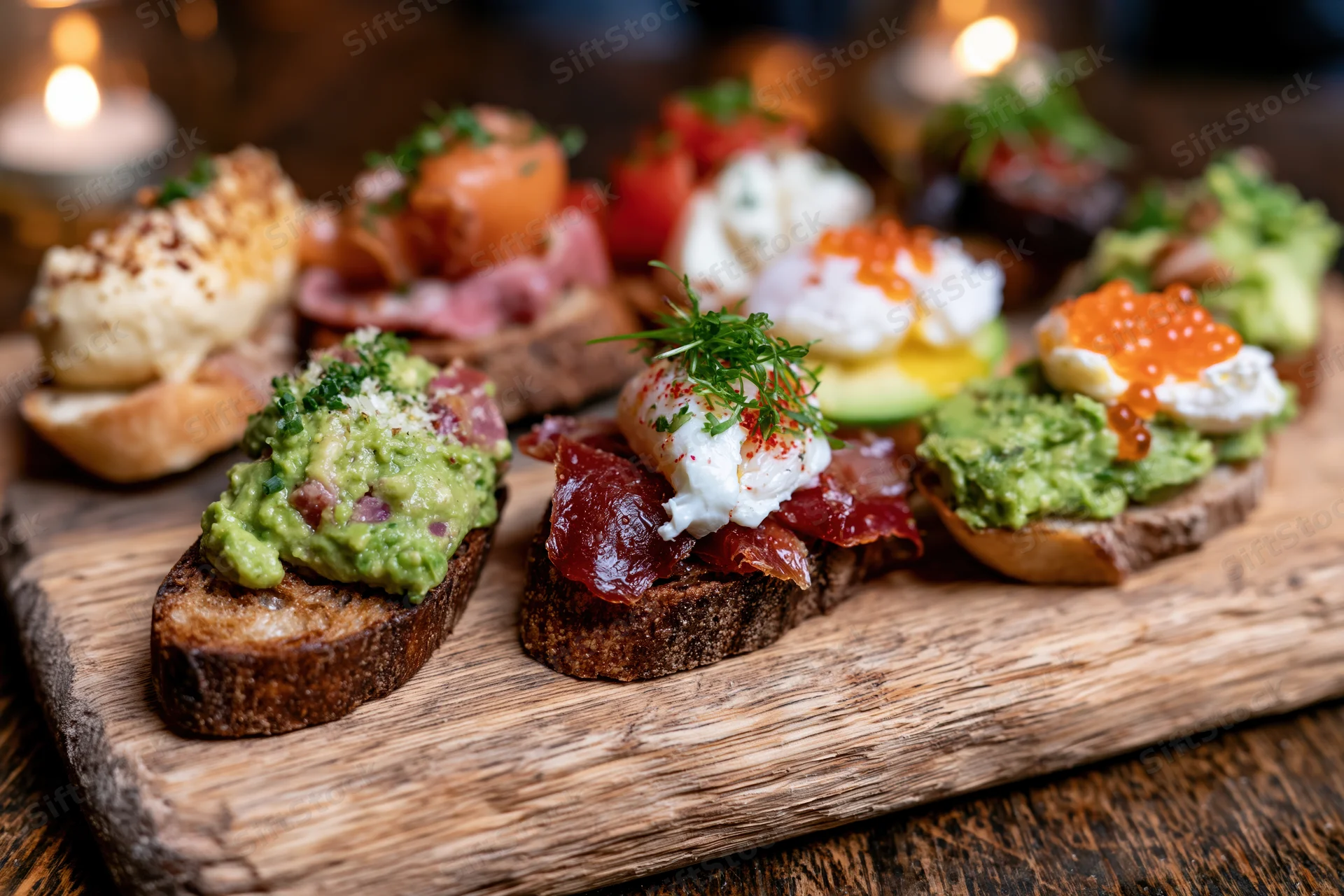 Assorted open-faced mini toasts with avocado, poached egg, salmon and caviar arranged on a wooden board under candlelight