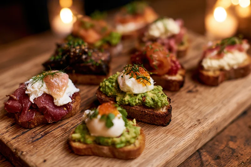 Assorted open-faced mini toasts and crostini arranged on a wooden cutting board with candles in the background
