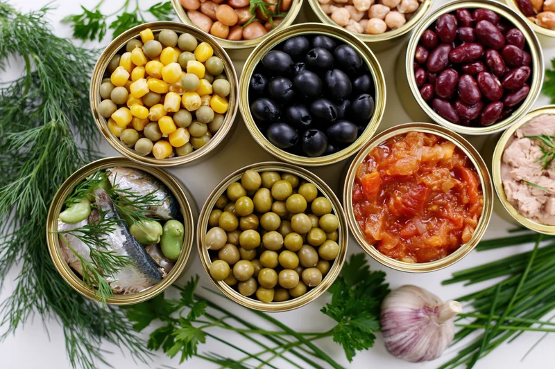 Open tin cans filled with assorted canned foods arranged with fresh herbs on white surface