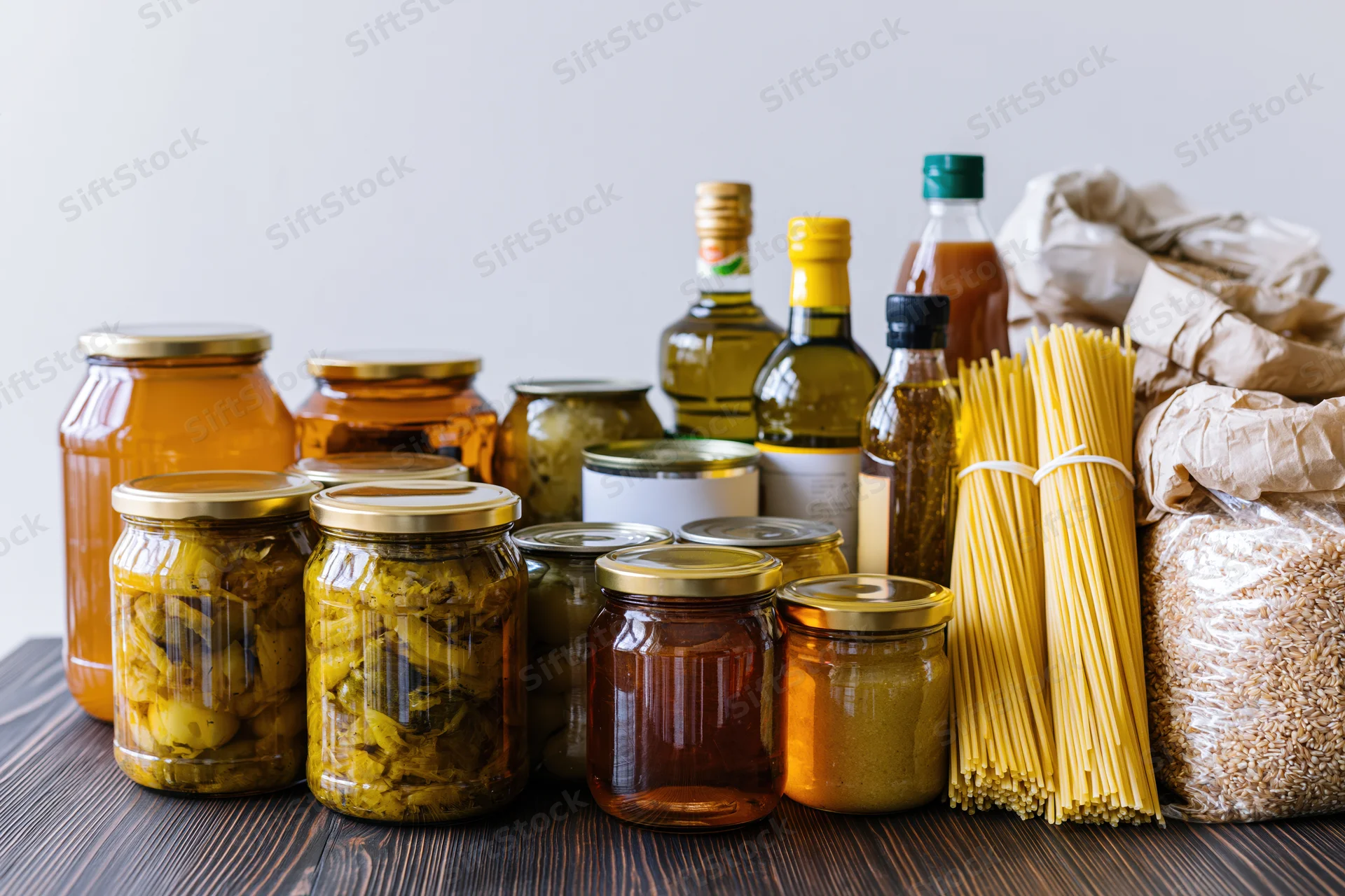 Assorted pantry staples arranged on dark wooden surface including jars of pickled vegetables, olive oil bottles, pasta, and rice bags