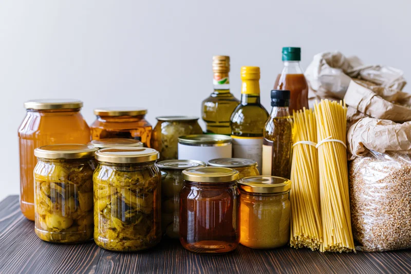 Assorted pantry staples arranged on dark wooden surface including jars of pickled vegetables, olive oil bottles, pasta, and rice bags