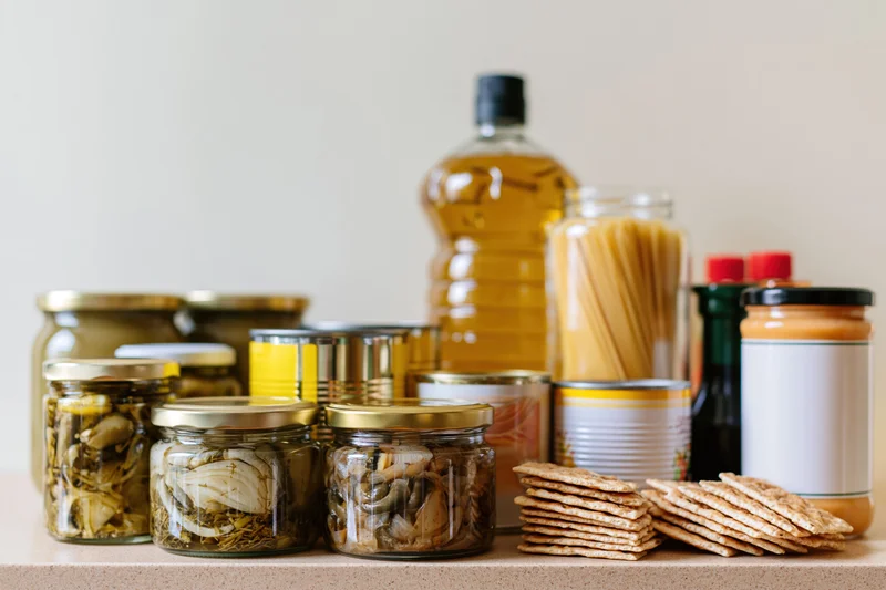 Assorted pantry staples arranged on a kitchen counter including jars of pickled vegetables, canned goods, pasta, crackers, oil, and peanut butter