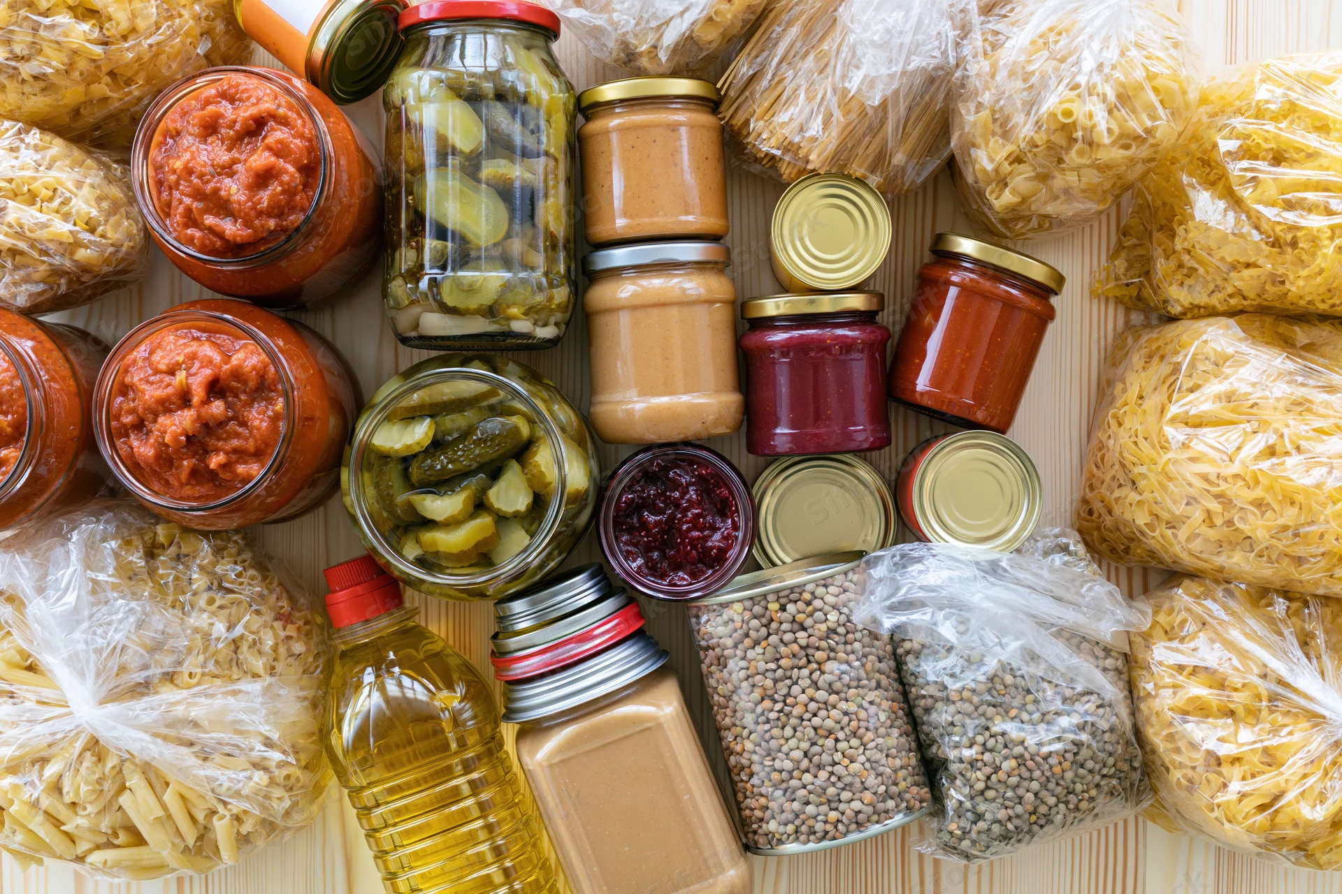 Assorted pantry staples including jars of pickles, tomato sauce, peanut butter, lentils, pasta, and oil arranged on a wooden surface