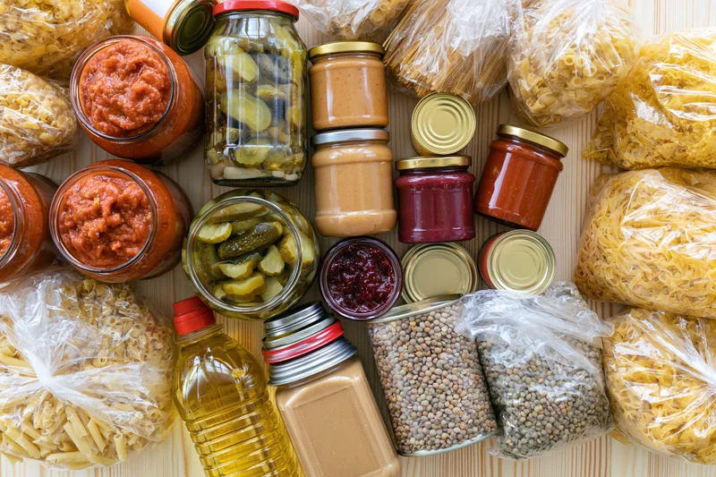 Assorted pantry staples including jars of pickles, tomato sauce, peanut butter, lentils, pasta, and oil arranged on a wooden surface