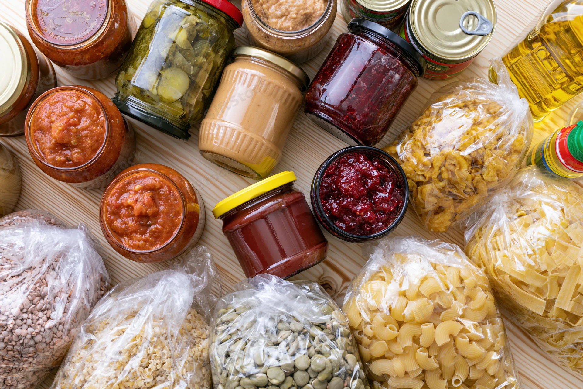 Assorted pantry staples including jars of sauce, jam, peanut butter, pickles, oil, and bags of pasta, beans, grains arranged on a wooden surface