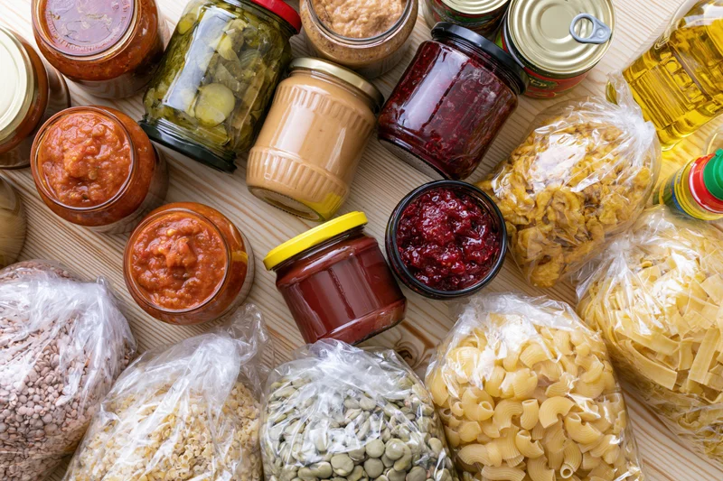 Assorted pantry staples including jars of sauce, jam, peanut butter, pickles, oil, and bags of pasta, beans, grains arranged on a wooden surface