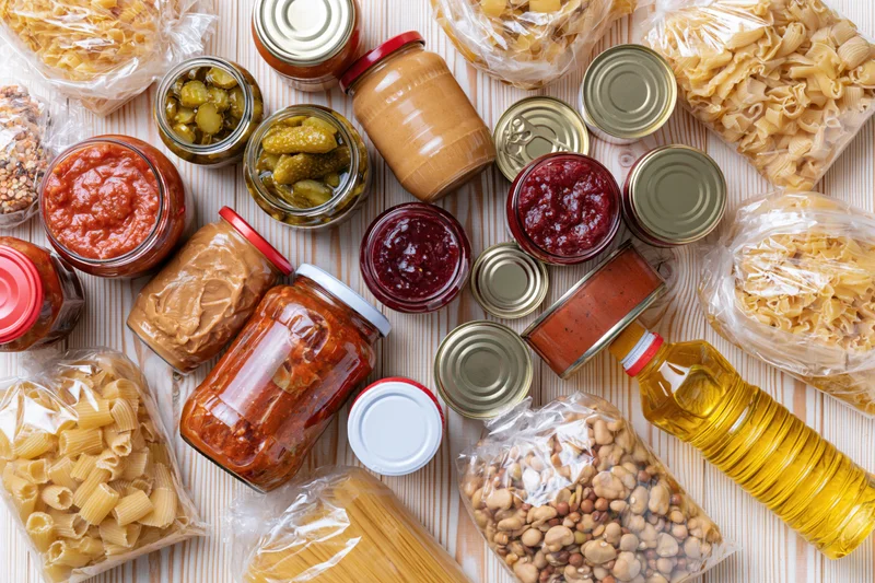 Assorted pantry staples including jars of sauce, peanut butter, jam, pickles, canned goods, pasta, beans, and oil arranged on a pale wooden surface