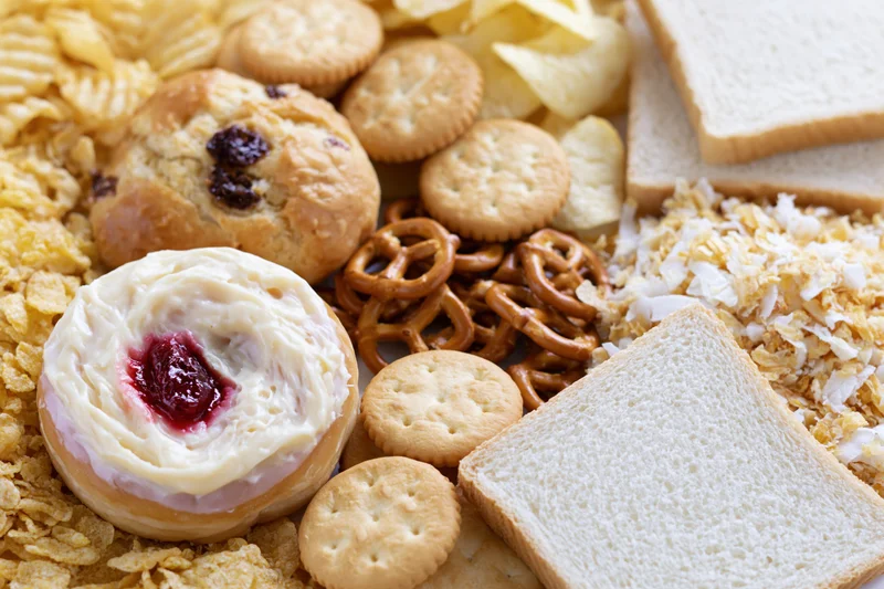 Assorted processed snack foods arranged together in a flat lay including crackers, chips, pretzels, donut, bread, and cereal