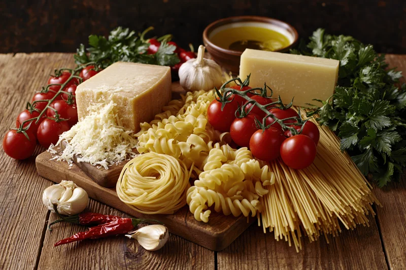 Assorted uncooked italian pasta varieties with cherry tomatoes cheese garlic and herbs arranged on a wooden board