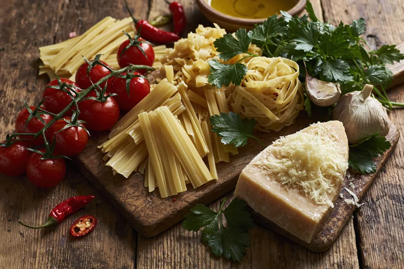 Uncooked italian pasta varieties with cherry tomatoes, parmesan cheese, garlic, parsley and chili on a wooden cutting board