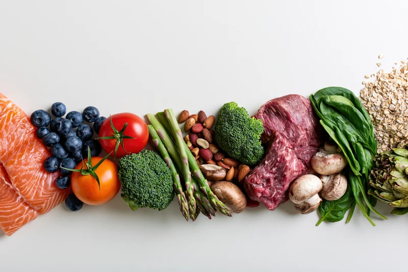 Assorted whole foods arranged in a row along the bottom edge of a white surface