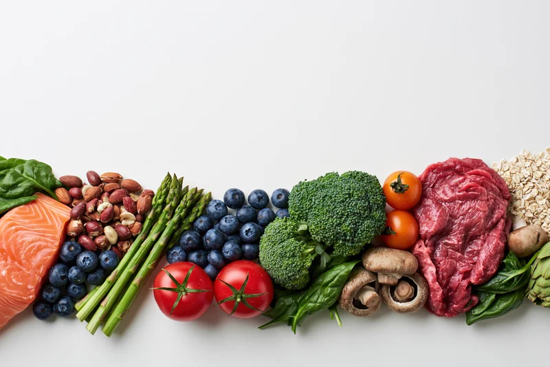 Assorted whole foods arranged in a row along the bottom edge of a white surface