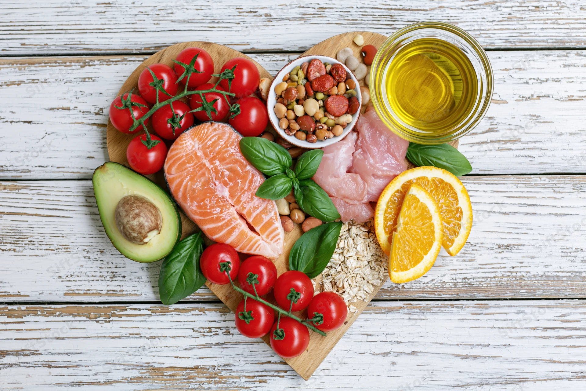 Heart-shaped arrangement of whole foods including salmon, avocado, tomatoes, orange, olive oil, beans, oats, and basil on a white wooden surface