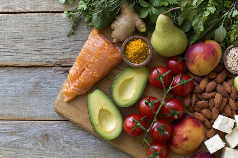 Salmon fillet, halved avocado, cherry tomatoes, pears, peaches, almonds, feta cheese, ginger, turmeric, herbs arranged on a wooden cutting board on a weathered wooden table