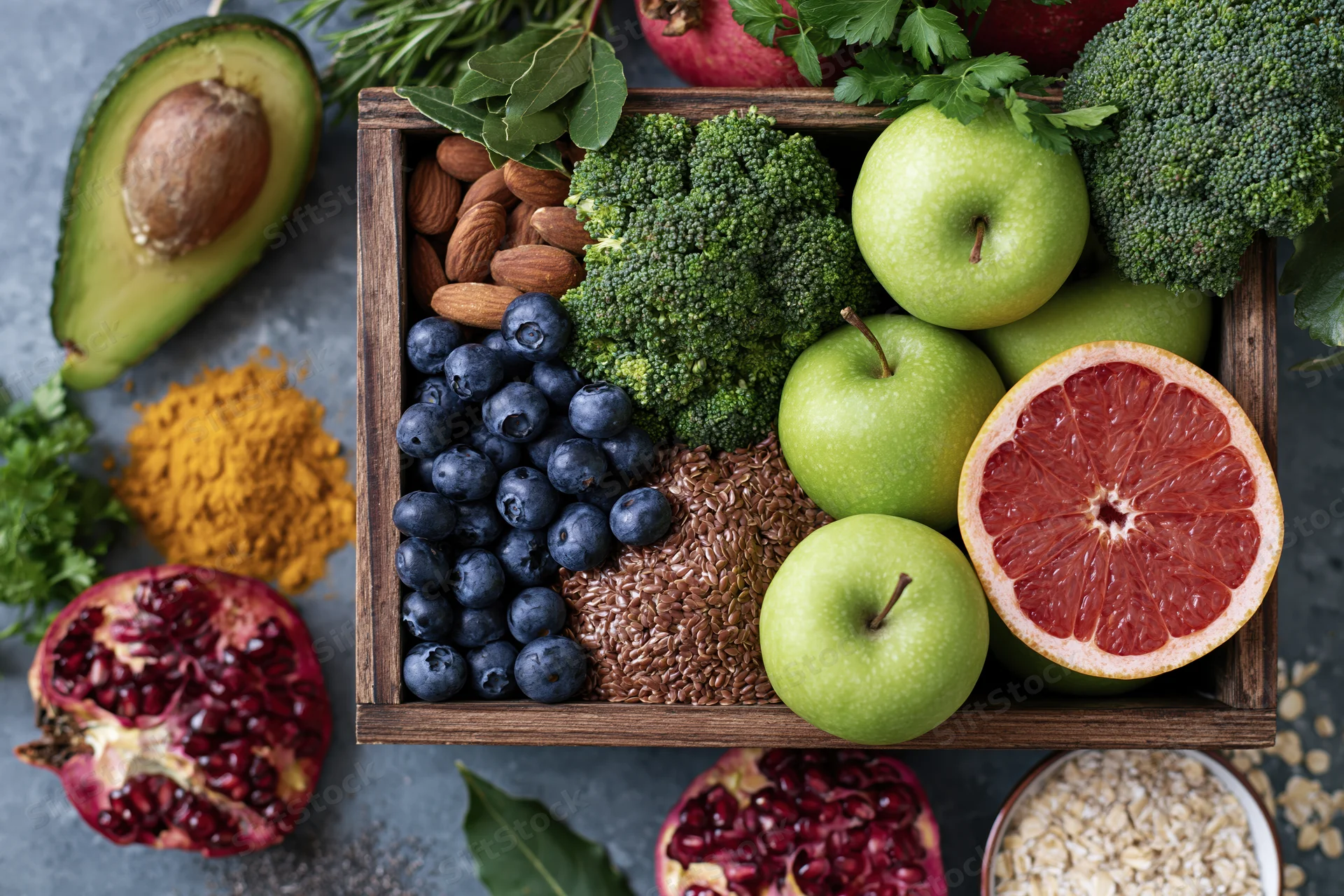 Fresh fruits and vegetables arranged in a wooden crate with nuts and seeds on a gray surface