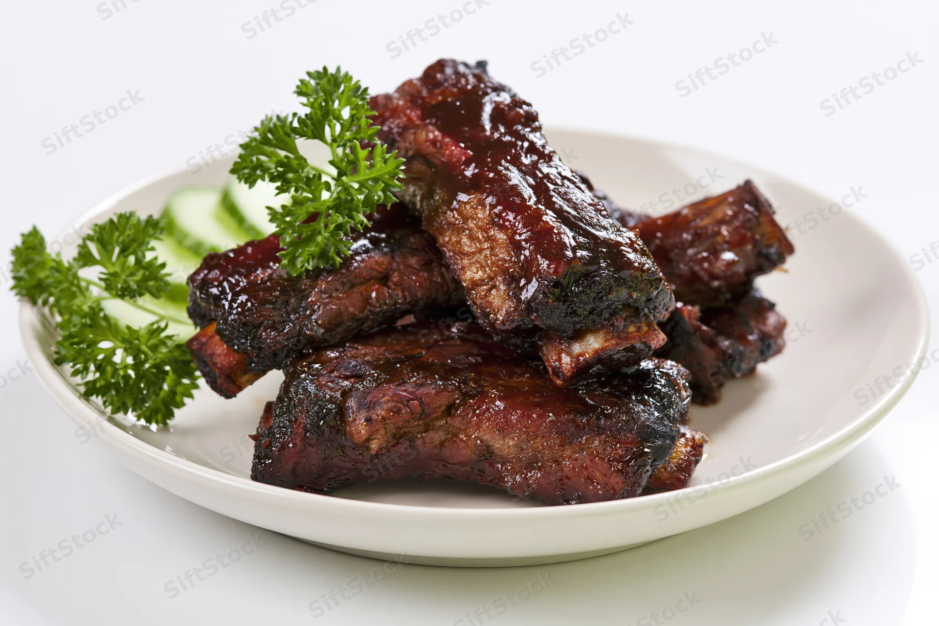 Glazed barbecue pork ribs stacked on a white plate with parsley and cucumber garnish