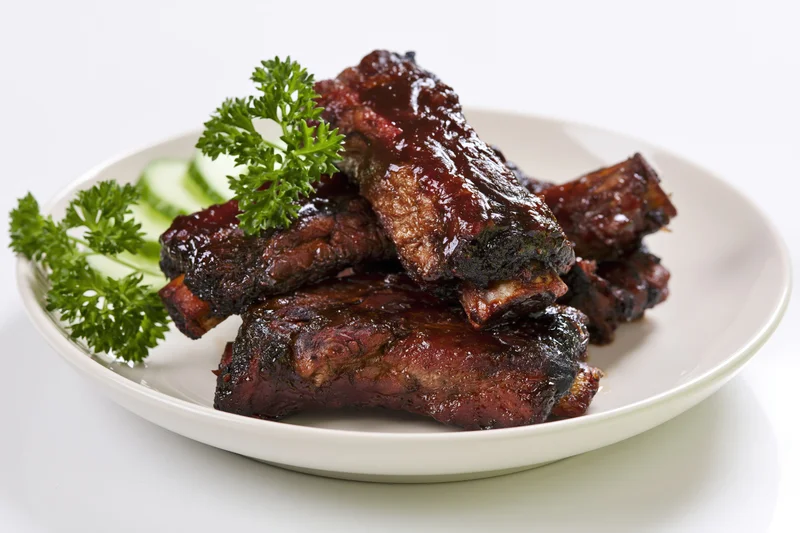 Glazed barbecue pork ribs stacked on a white plate with parsley and cucumber garnish