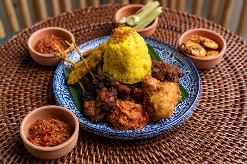 Balinese nasi kuning feast served on a blue patterned plate with satay and sambal on a woven rattan mat