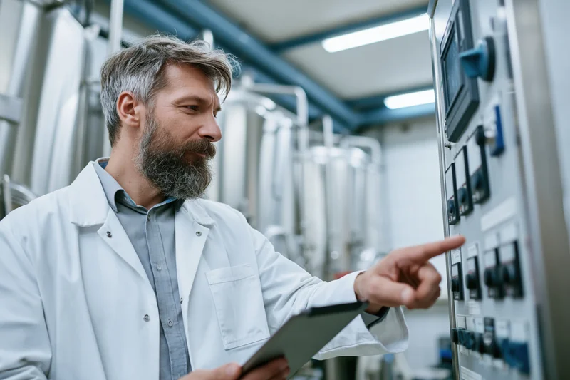 Bearded male technician in white lab coat holding tablet and pointing at control panel in factory