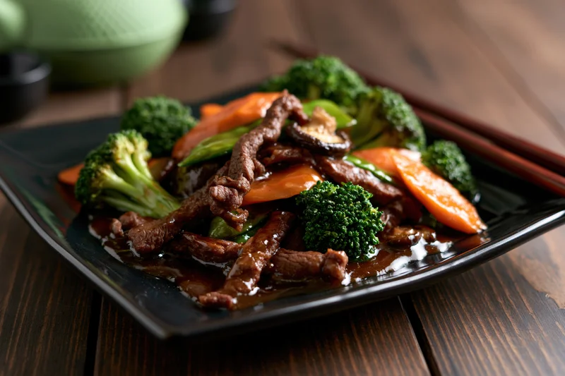 Beef and vegetable stir-fry served on a square black ceramic plate with chopsticks on a wooden table