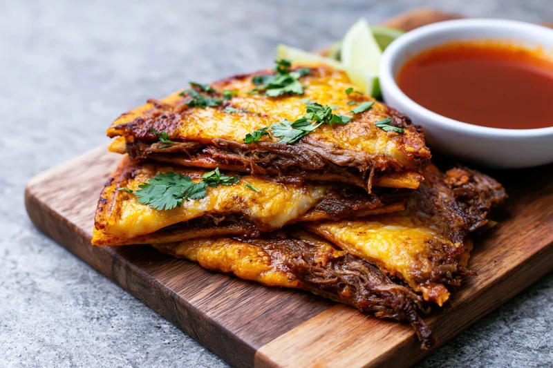 Birria quesatacos stacked on a wooden cutting board with salsa and cilantro garnish