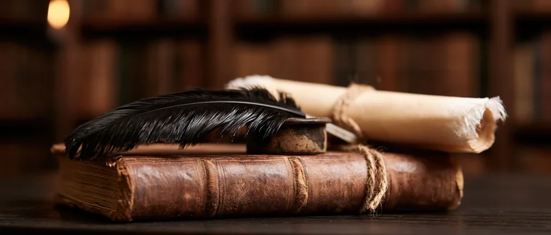 Black feather quill resting in inkwell on leather-bound book with rolled parchment scroll, in dimly lit library setting