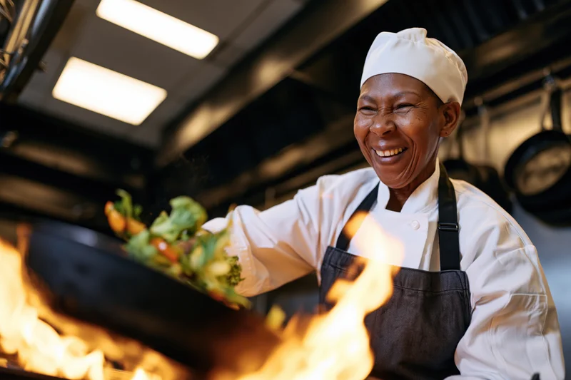 Black female chef smiling while tossing vegetables in a flaming pan in a commercial kitchen