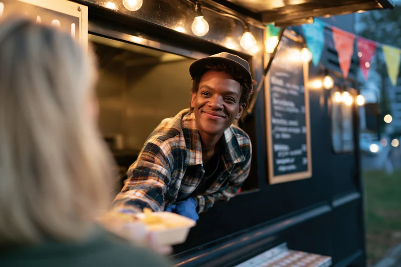 Young black man smiling while serving food from a food truck window at night
