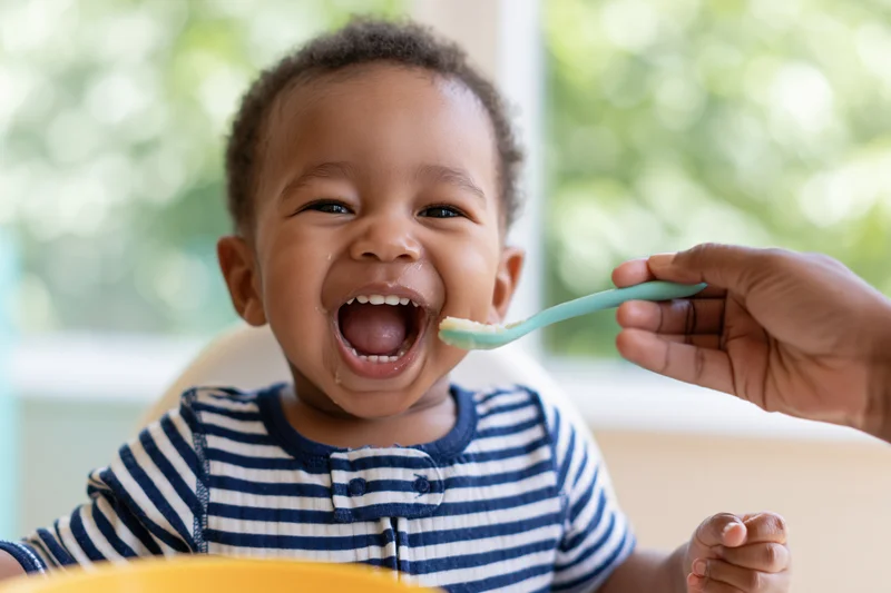 Toddler boy laughing with open mouth as hand feeds him puree with spoon in highchair