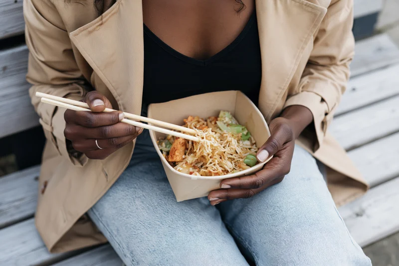 Woman eating takeout noodles with chopsticks while sitting on a wooden outdoor bench