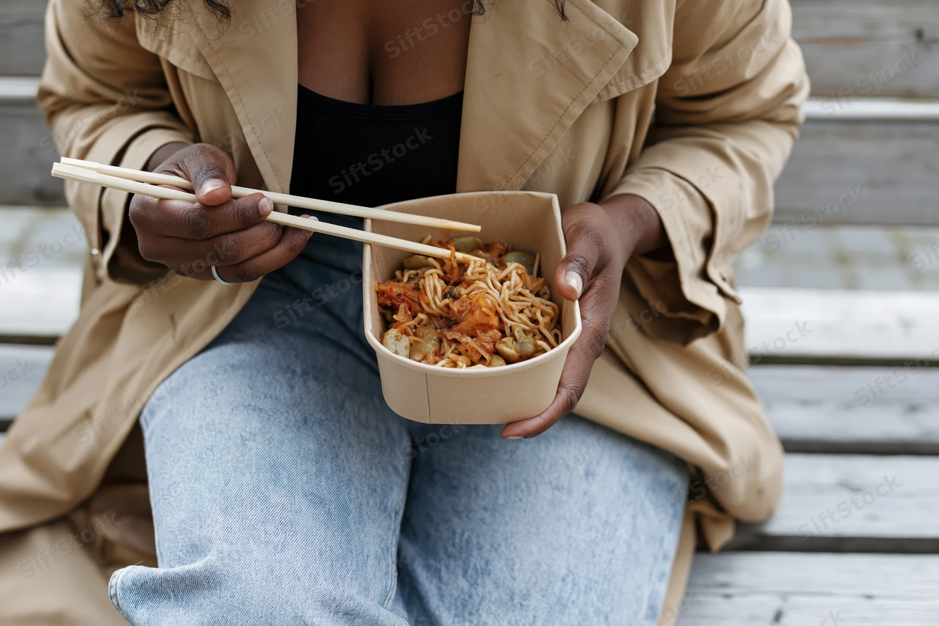 Woman eating noodles from a takeout bowl with chopsticks while sitting on a wooden bench